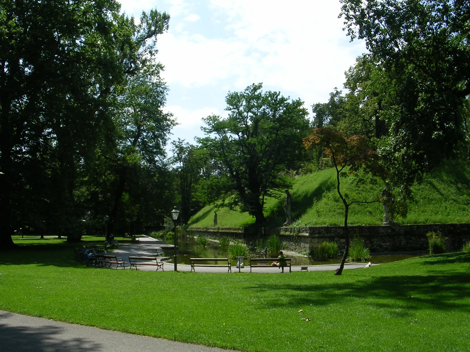 Weite Wiese im Grazer Stadtpark mit Bänken, Laterne, Teich und begrüntem Wall unter großen Laubbäumen