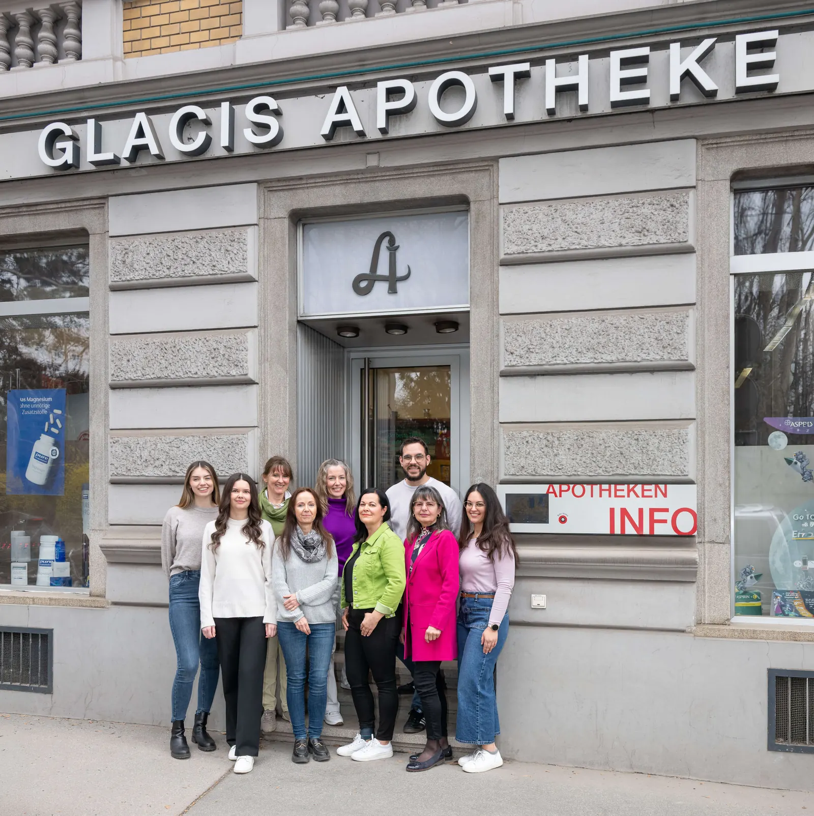 Das Team der Glacis-Apotheke steht vor dem Eingang des Geschäfts mit dem Schriftzug „Glacis Apotheke“ und dem Apotheken-A